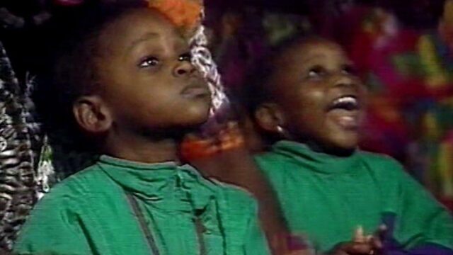 A video still shows two children dressed in green, one looks up smiling in excitement and the other looks up concernedly.